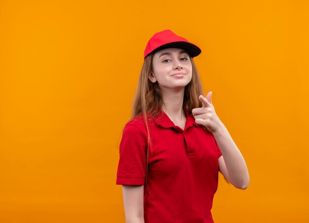 Zelfverzekerd jong leveringsmeisje in rood uniform doet je gebaar op geïsoleerde oranje ruimte met exemplaarruimte
