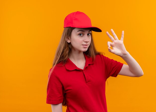 Zelfverzekerd jong leveringsmeisje in rood uniform doet ok teken op geïsoleerde oranje ruimte met exemplaarruimte