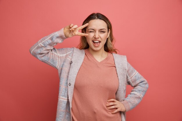 Zelfverzekerd jong meisje die hand op taille houden die v-sign-symbool dichtbij oog knipogen tonen