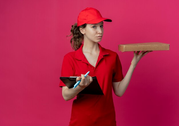 Zelfverzekerd jong mooi bezorgmeisje in rood uniform en pet met pizzapakketpen en klembord geïsoleerd op karmozijnrode achtergrond met kopie ruimte
