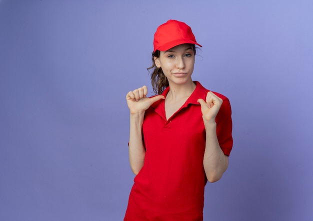 Zelfverzekerd jong mooi leveringsmeisje die rood uniform en GLB dragen die naar zichzelf richten geïsoleerd op paarse achtergrond met exemplaarruimte