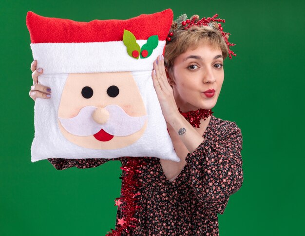 Zelfverzekerd jong mooi meisje met de kroon van het kersthoofd en klatergoudslinger om de nek met het kussen van de kerstman en raakt het hoofd ermee aan met getuite lippen geïsoleerd op een groene muur