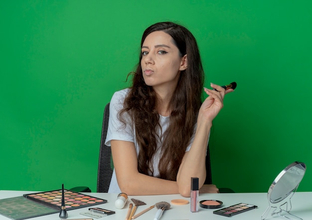 Zelfverzekerd jong mooi meisje zittend aan de make-up tafel met make-up tools houden blozen borstel hand zetten tafel geïsoleerd op groene achtergrond