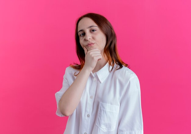 Zelfverzekerd jong roodharigemeisje die hand onder kin zetten die op roze achtergrond wordt geïsoleerd