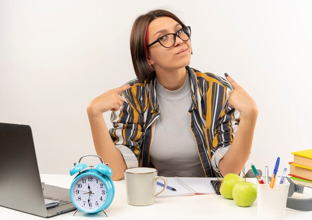 Zelfverzekerd jong studentenmeisje die glazen dragen die bij bureau zitten die op zichzelf richten op wit wordt geïsoleerd