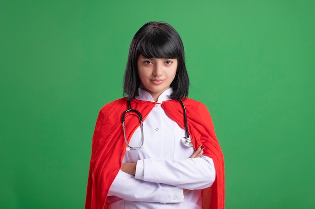 Zelfverzekerd jong superheromeisje die stethoscoop met medisch kleed en mantel dragen die handen kruisen die op groen worden geïsoleerd