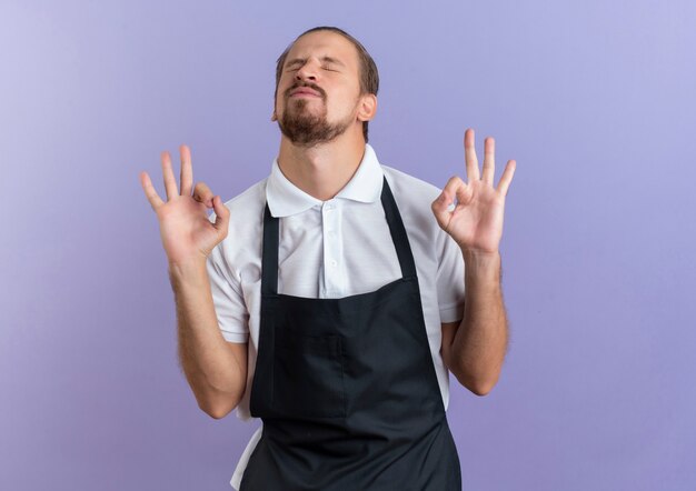 Zelfverzekerd jonge knappe kapper dragen uniform doen ok tekenen met gesloten ogen geïsoleerd op paars met kopie ruimte