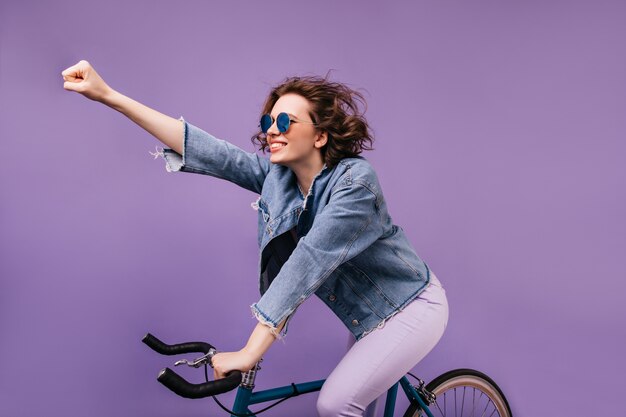 Zelfverzekerd meisje in spijkerjasje rijden op de fiets en zwaaiende hand. Indoor foto van geïnspireerde jonge dame in glazen zittend op de fiets.