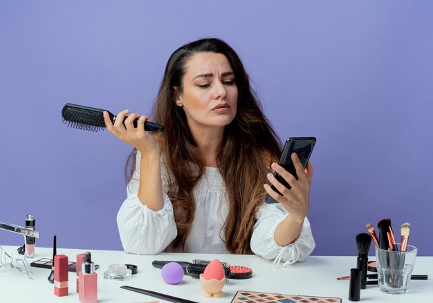 Zelfverzekerd mooi meisje zit aan tafel met make-up tools houdt haar kam kijken naar telefoon geïsoleerd op paarse muur