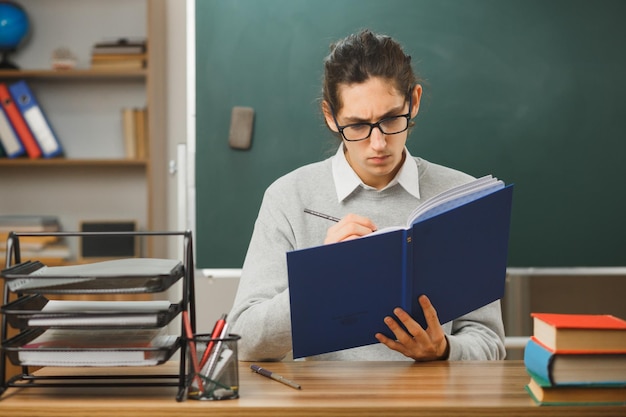 zelfverzekerd schrijven op notitieboekje jonge mannelijke leraar zit aan bureau met schoolhulpmiddelen in de klas