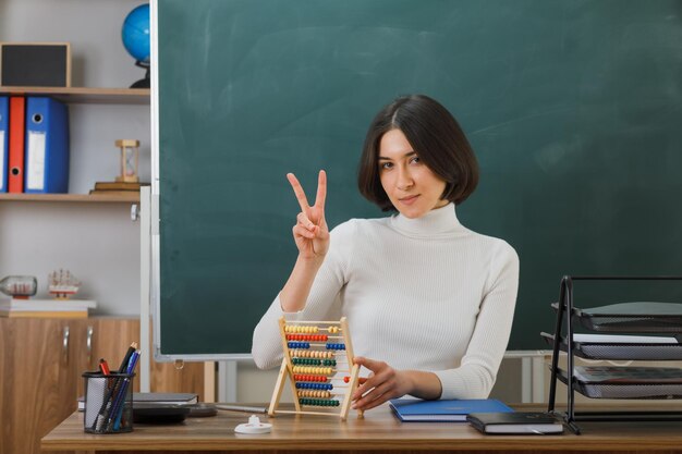 zelfverzekerd tonen van vredesgebaar jonge vrouwelijke leraar met telraam zittend aan bureau met schoolhulpmiddelen in de klas