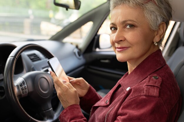 Zelfverzekerd volwassen zakenvrouw in stijlvolle jas chats met haar smartphone in de auto