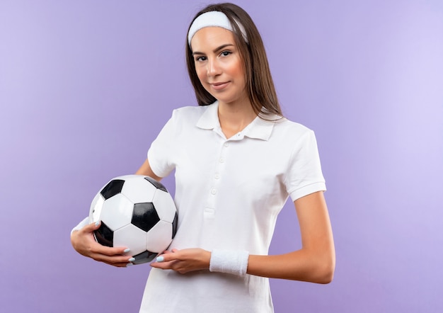 Zelfverzekerd vrij sportief meisje met hoofdband en polsbandje met voetbal geïsoleerd op paarse muur