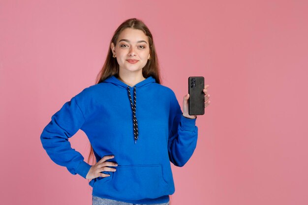 Zelfverzekerd vrouwelijk model dat één hand op de taille houdt en moderne smartphone demonstreert op camera