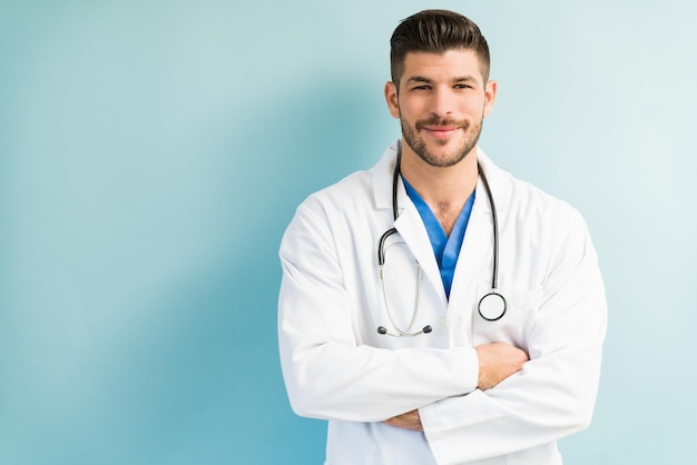 Zelfverzekerde aantrekkelijke mannelijke arts die een witte laboratoriumjas draagt terwijl hij staat met gekruiste armen tegen een turkooizen achtergrond