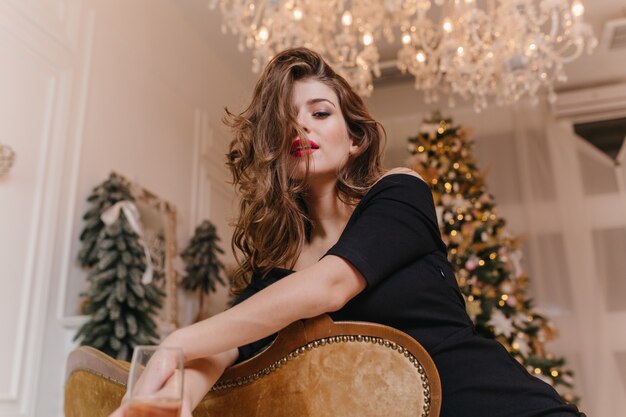 Zelfverzekerde aantrekkelijke vrouw poseren na nieuwjaarsfeest, zittend op bruine bank. Indoor portret van knappe blanke meisje ontspannen thuis voor kerstboom.