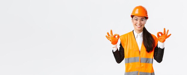 Zelfverzekerde Aziatische vrouwelijke bouwingenieur-ondernemingsmanager die een goed gebaar toont na het opzetten van een veiligheidshelm, bril en handschoenen voordat hij een gevaarlijk gebied betreedt op een witte achtergrond