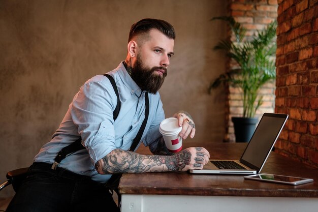 Zelfverzekerde bebaarde man leunt op de tafel. Hij draagt overhemd en jarretel. Hij heeft tatoeages op zijn armen en nek. De mens werkt aan zijn laptop. kantoor op de achtergrond.