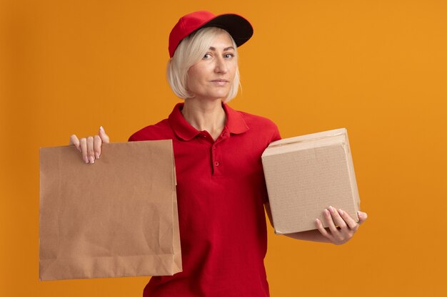 Zelfverzekerde blonde bezorger van middelbare leeftijd in rood uniform en pet met kartonnen doos en papieren pakket