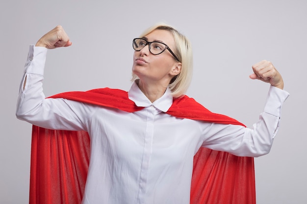 Zelfverzekerde blonde superheld vrouw van middelbare leeftijd in rode cape met een bril die een sterk gebaar doet en omhoog kijkt geïsoleerd op een witte muur