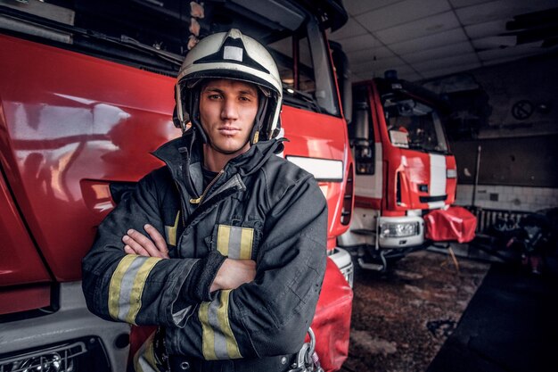 Zelfverzekerde brandweerman met een beschermend uniform dat naast een brandweerwagen staat in een garage van een brandweer, gekruiste armen en kijkend naar een camera