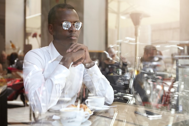 Zelfverzekerde doordachte jonge Afro-Amerikaanse zakenman in stijlvolle brillen handen omklemde zittend in de coffeeshop met mok op tafel, kwam na te denken over zakelijke problemen over kopje cappuccino