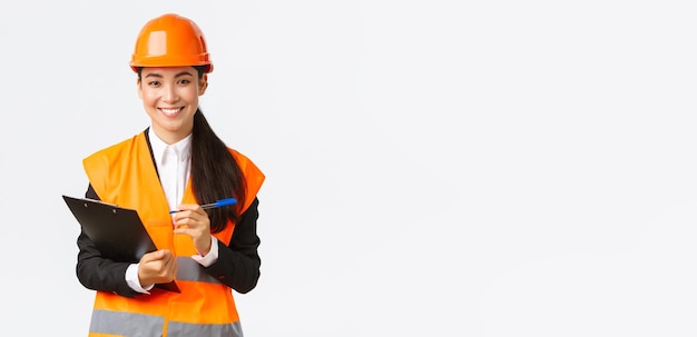Zelfverzekerde glimlachende vrouwelijke Aziatische bouwingenieur industriële vrouw in veiligheidshelm bezoek bouwgebied voor inspectie notities op klembord opschrijven en tevreden witte achtergrond kijken