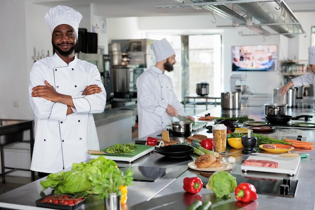 Zelfverzekerde hoofdkok die in de professionele keuken van het restaurant staat met gekruiste armen terwijl hij naar de camera glimlacht. Souschef draagt kookuniform terwijl hij ingrediënten voorbereidt voor het avondeten.