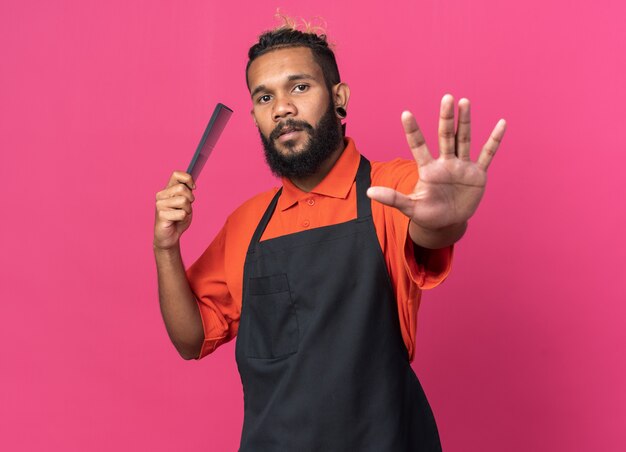 Zelfverzekerde jonge Afro-Amerikaanse mannelijke kapper die een uniforme kam draagt die een stopgebaar doet