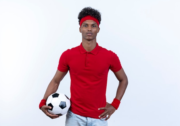 Zelfverzekerde jonge Afro-Amerikaanse sportieve man met haedband en polsbandje houden bal en hand op heup zetten geïsoleerd op een witte achtergrond