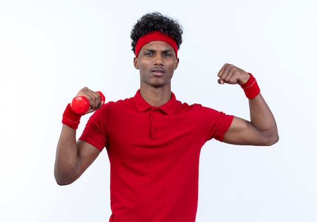 Zelfverzekerde jonge Afro-Amerikaanse sportieve man met haedband en polsbandje houdt bumbbell en doet sterk gebaar geïsoleerd op witte achtergrond