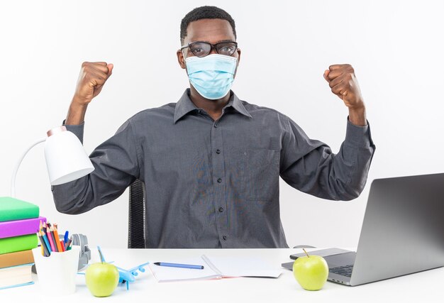 Zelfverzekerde jonge Afro-Amerikaanse student in optische bril met een medisch masker zittend aan een bureau met schoolhulpmiddelen die vuisten op een witte muur houden