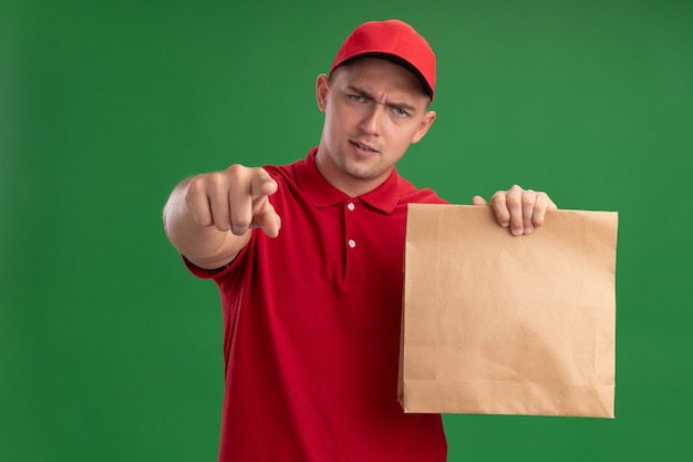 Zelfverzekerde jonge bezorger die uniform en pet draagt en papier voedselpakket houdt dat je gebaar toont dat op groene muur wordt geïsoleerd