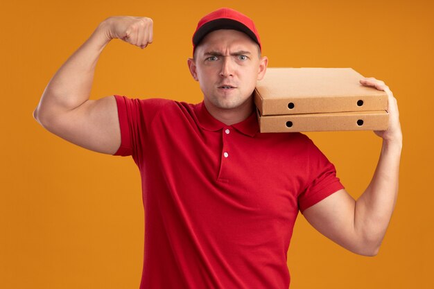 Zelfverzekerde jonge bezorger die uniform met pet draagt die pizzadozen op schouder houdt die sterk gebaar tonen dat op oranje muur wordt geïsoleerd