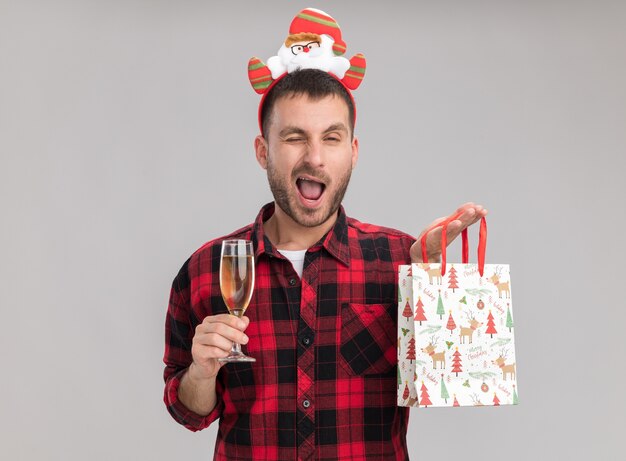 Zelfverzekerde jonge blanke man met kerst hoofdband houden kerst cadeau zak en glas champagne kijken camera knipogen geïsoleerd op een witte achtergrond met kopie ruimte