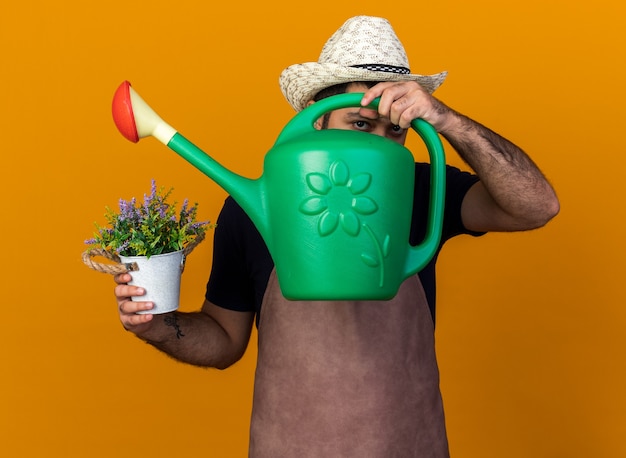 Zelfverzekerde jonge blanke mannelijke tuinman met een tuinhoed met bloempot en kijkend door een gieter geïsoleerd op een oranje muur met kopieerruimte