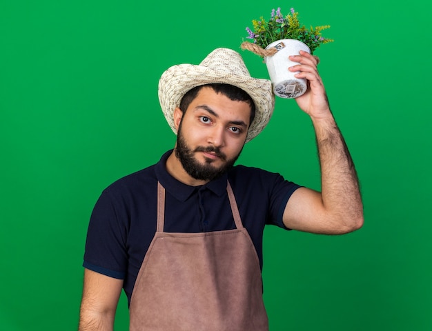 Gratis foto zelfverzekerde jonge blanke mannelijke tuinman met een tuinhoed met een bloempot boven het hoofd geïsoleerd op een groene muur met kopieerruimte