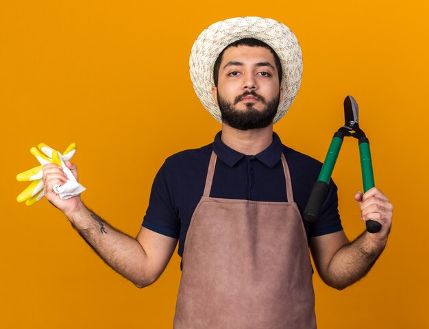 zelfverzekerde jonge blanke mannelijke tuinman tuinieren hoed bedrijf tuinieren schaar en handschoenen geïsoleerd op oranje muur met kopie ruimte