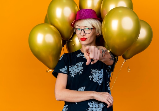 Zelfverzekerde jonge blonde feestvrouw met feestmuts en bril staan voor ballonnen kijken en wijzen naar voorzijde geïsoleerd op oranje muur