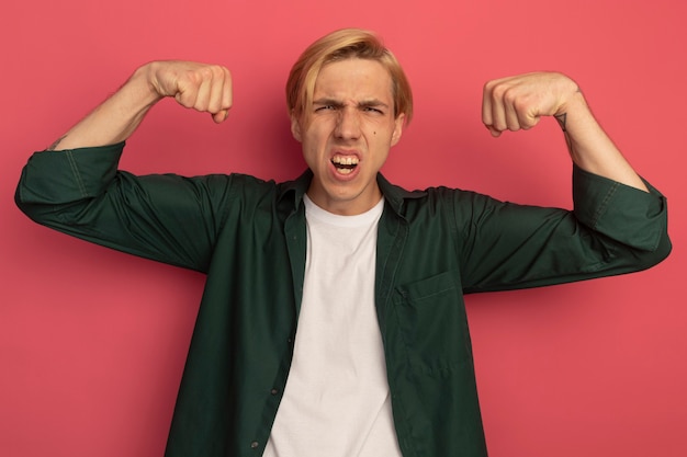 Zelfverzekerde jonge blonde kerel die groen t-shirt draagt dat sterk gebaar doet dat op roze wordt geïsoleerd