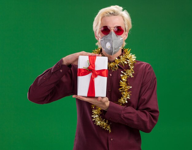 Zelfverzekerde jonge blonde man met bril en beschermend masker met klatergoud slinger rond nek houden geschenkpakket geïsoleerd op groene muur