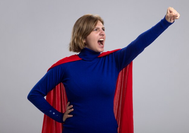 Zelfverzekerde jonge blonde supervrouw in rode cape die vuist opheft opstaand in superman pose kijkend naar kant geïsoleerd op een witte muur