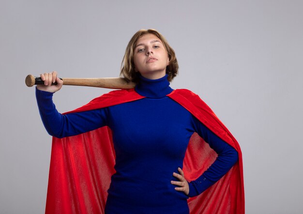Zelfverzekerde jonge blonde supervrouw in rode cape met honkbalknuppel die hand op taille houdt die voorzijde bekijkt die op witte muur wordt geïsoleerd