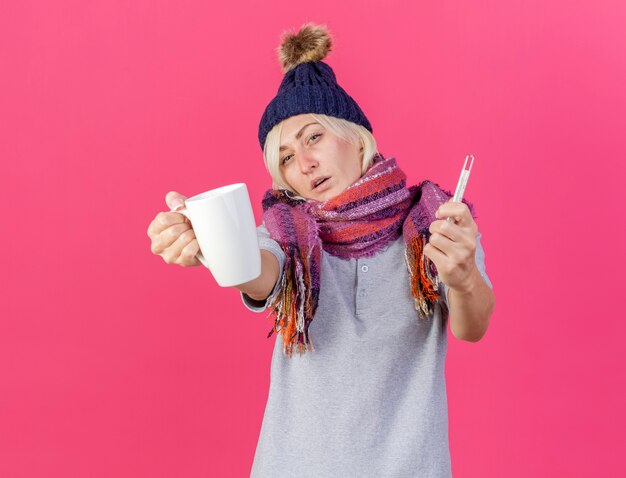 Zelfverzekerde jonge blonde zieke Slavische vrouw met muts en sjaal houdt beker en thermometer geïsoleerd op roze muur met kopie ruimte