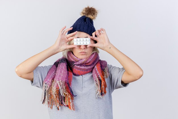 Zelfverzekerde jonge blonde zieke Slavische vrouw met winter muts en sjaal houdt pakje medische pillen voor ogen geïsoleerd op een witte muur met kopie ruimte