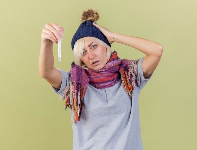 Zelfverzekerde jonge blonde zieke slavische vrouw met winter muts en sjaal legt hand op het hoofd en houdt thermometer geïsoleerd op olijfgroene muur met kopie ruimte