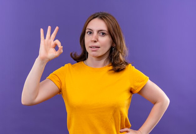 Zelfverzekerde jonge casual vrouw doet ok teken met hand op taille op geïsoleerde paarse ruimte