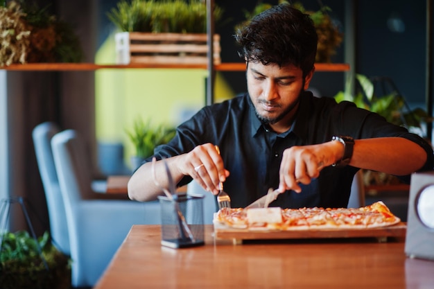 Zelfverzekerde jonge indiase man in zwart shirt zit bij pizzeria met pizza