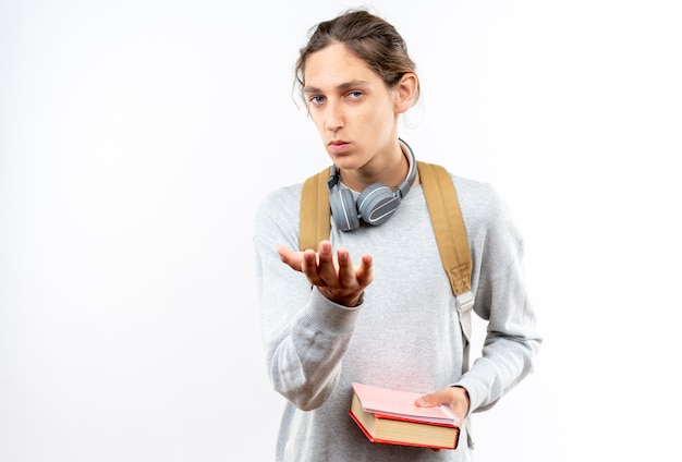 Zelfverzekerde jonge kerel student draagt rugzak met koptelefoon op nek met boeken geïsoleerd op een witte muur