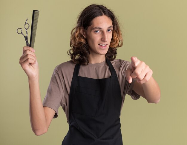 Zelfverzekerde jonge knappe kapper met een uniforme schaar en kam die kijkt en wijst naar de voorkant geïsoleerd op een olijfgroene muur